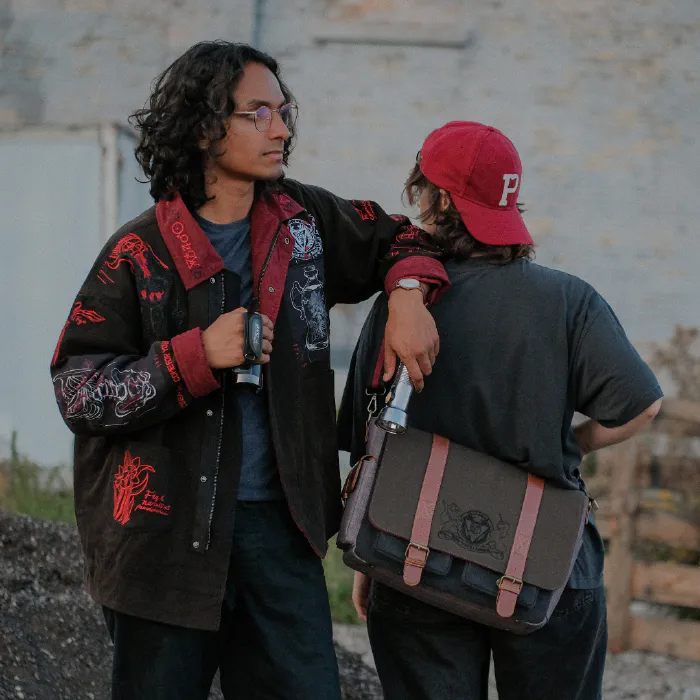 Two people model the Knock Thrice The Magnus Protocol merchandise. This picture features an embroidered jacket and a custom OIAR messenger bag.