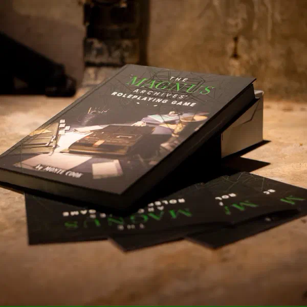 The Magnus Archives Roleplaying Game hardcover book lying on a stone floor with promotional postcards fanned out beneath it.